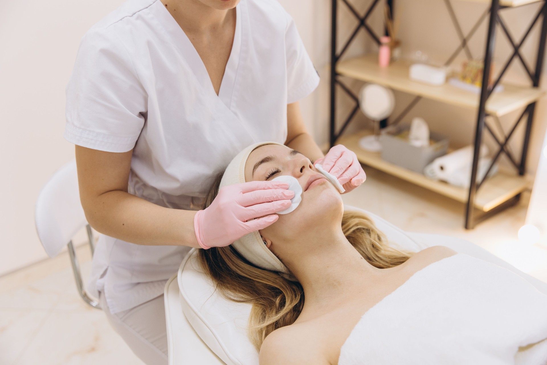 Beautician wearing pink gloves cleaning face of young blond woman lying on white bed with cotton pads in beauty salon